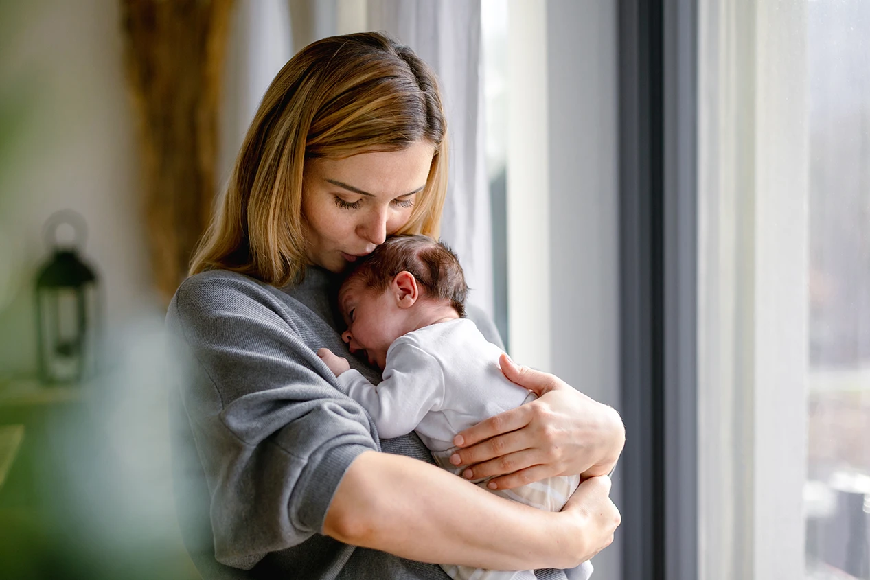 A mother gently holding and kissing her sleeping newborn by a window